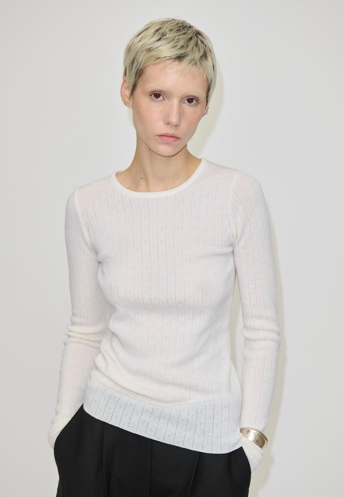 A person wearing the Herskind ApS Camb Knit Blouse, a long-sleeved, white merino wool ribbed knit top with pointelle detailing.