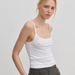 A person wearing a slim fit, white scoop-neck Poppy Top by Herskind ApS with thin straps and dark grey trousers.