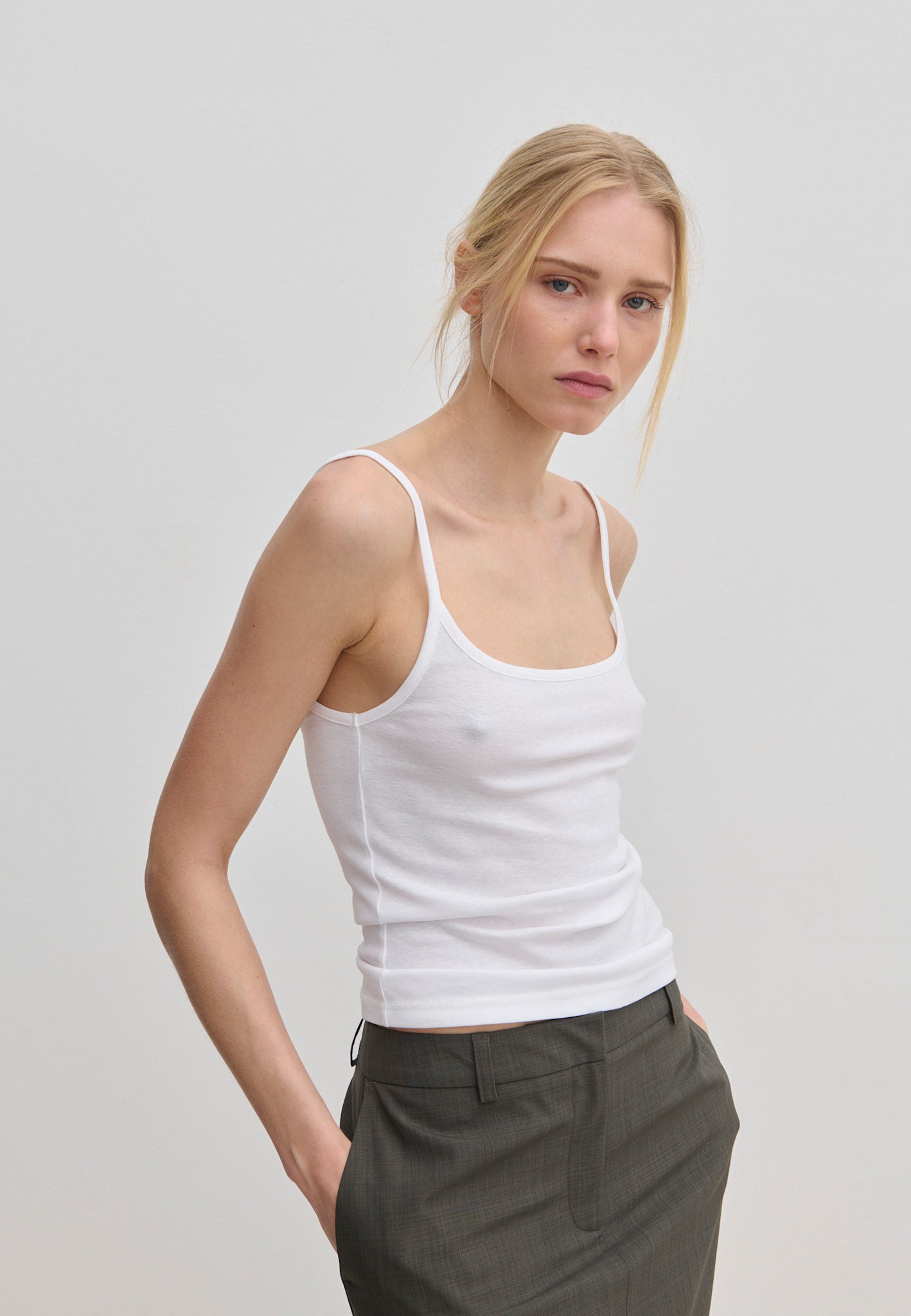 A person wearing a slim fit, white scoop-neck Poppy Top by Herskind ApS with thin straps and dark grey trousers.