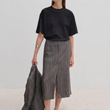 A person wears a boxy-fit black Larsson T-shirt by Herskind ApS and a grey plaid midi skirt with a front slit, holding a matching blazer in one hand.