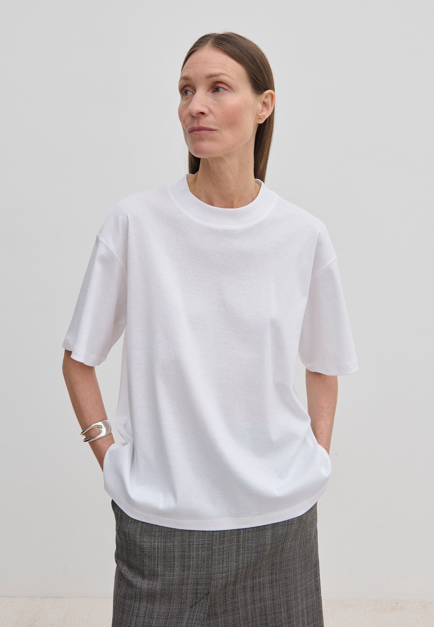 A person wears an oversized, organic cotton Herskind ApS Larsson T-shirt with a high neckline and grey patterned trousers.