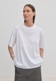 A person wears an oversized, organic cotton Herskind ApS Larsson T-shirt with a high neckline and grey patterned trousers.