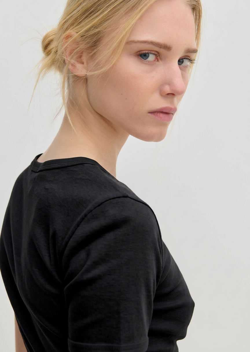 A person wearing a plain black, slim fit crew-neck River T-Shirt by Herskind ApS looks over their shoulder toward the camera.