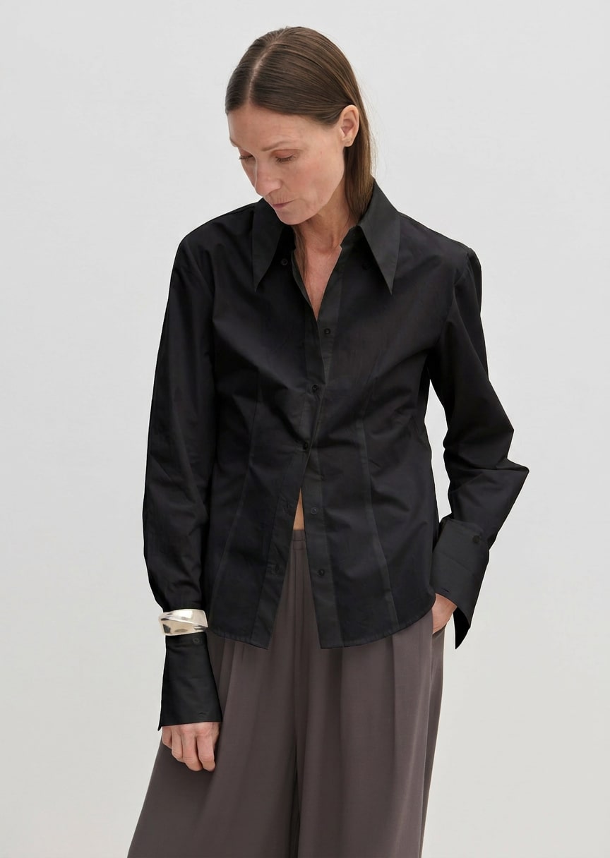 A slim-fit, long-sleeved black Vivienne Shirt by Herskind ApS with a pointed collar and wide cuffs, worn over grey trousers.