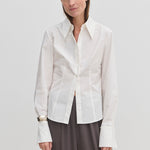 A person wearing a Vivienne Shirt by Herskind ApS, a slim fit, white, long-sleeved button-down with a pointed collar and wide cuffs, paired with dark grey trousers.