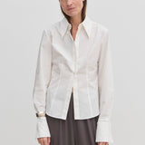 A person wearing a Vivienne Shirt by Herskind ApS, a slim fit, white, long-sleeved button-down with a pointed collar and wide cuffs, paired with dark grey trousers.