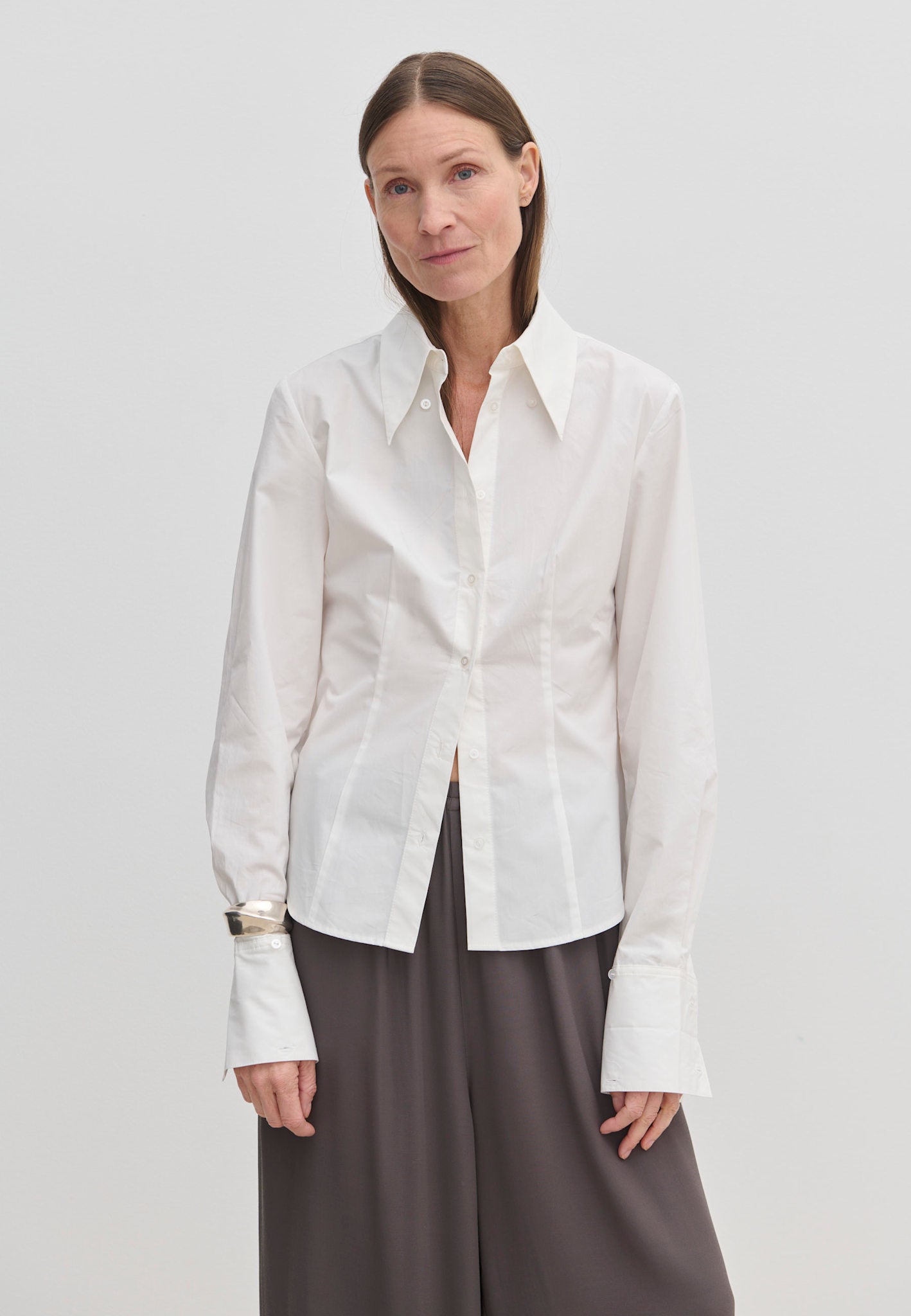 A person wearing a Vivienne Shirt by Herskind ApS, a slim fit, white, long-sleeved button-down with a pointed collar and wide cuffs, paired with dark grey trousers.