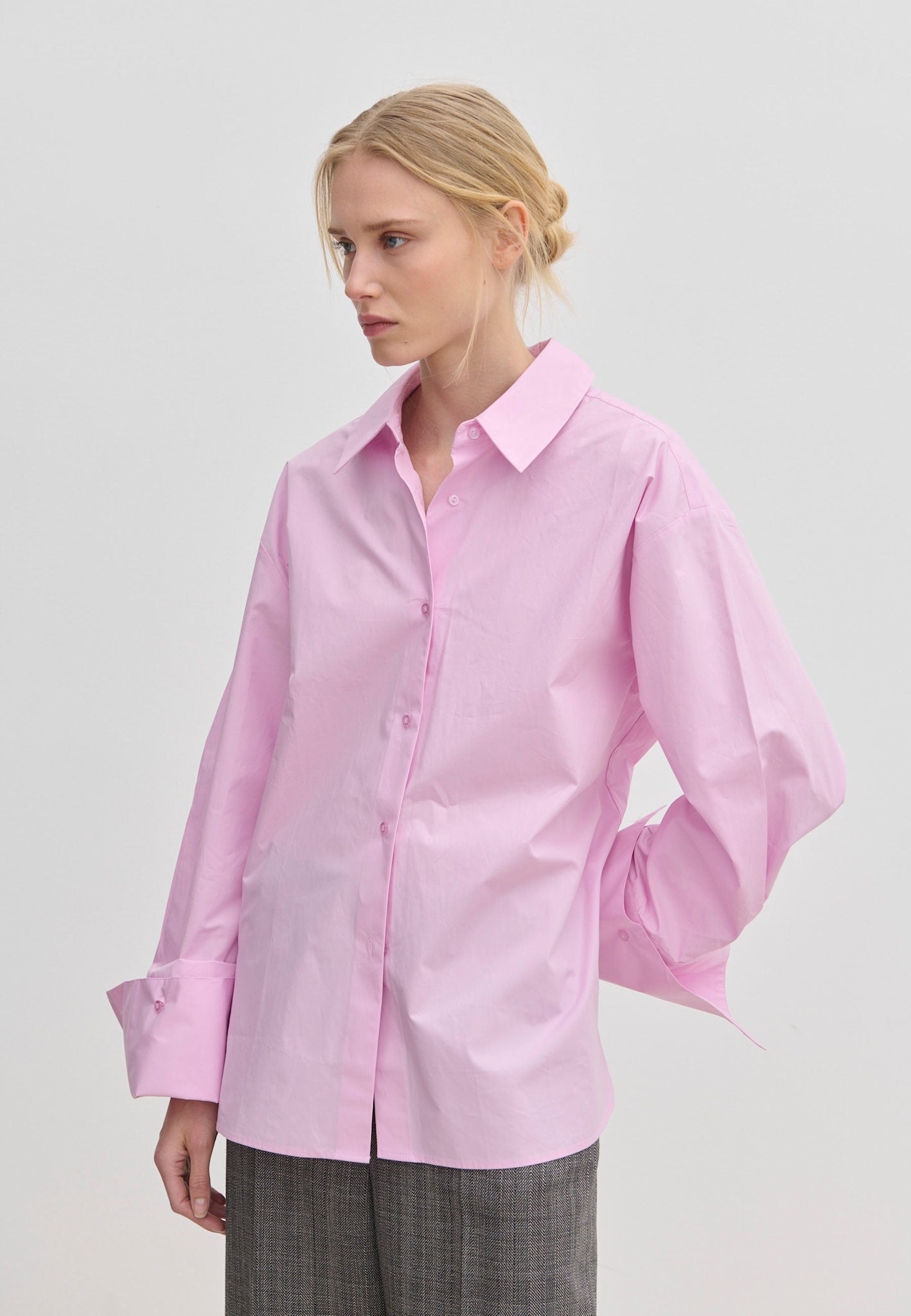 A woman wears an oversized light pink cotton Inga Shirt by Herskind ApS with wide, folded cuffs, paired with grey trousers.