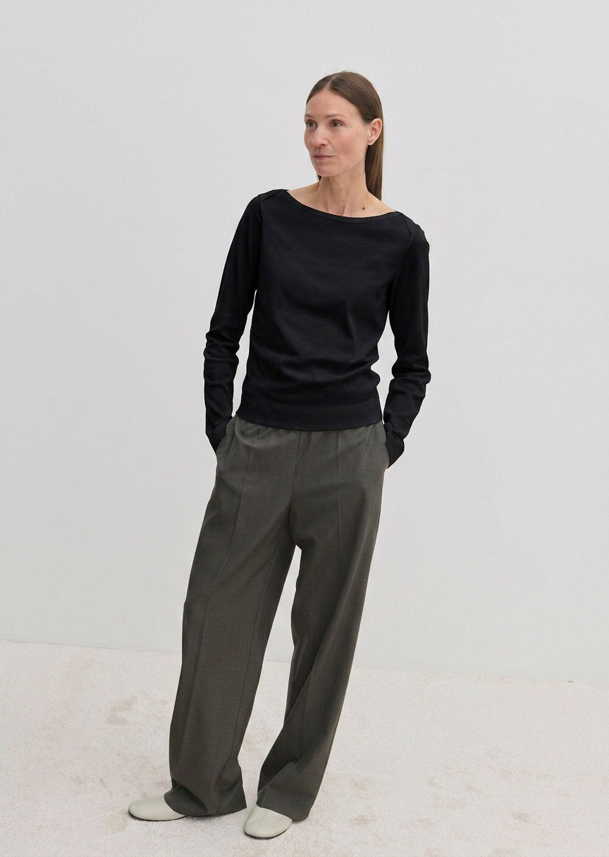 A woman wears a black, long-sleeved Emma Blouse by Herskind ApS with a boat neckline, paired with loose-fitting grey trousers.