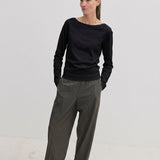 A woman wears a black, long-sleeved Emma Blouse by Herskind ApS with a boat neckline, paired with loose-fitting grey trousers.