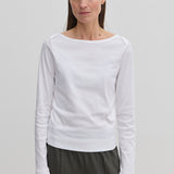 A woman wearing an Emma Blouse by Herskind ApS featuring a long-sleeved boat neckline and dark trousers.