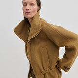 A woman wears a tan, funnel neck Oscar Wool Jacket by Herskind ApS with a zip front and a cinched waist.