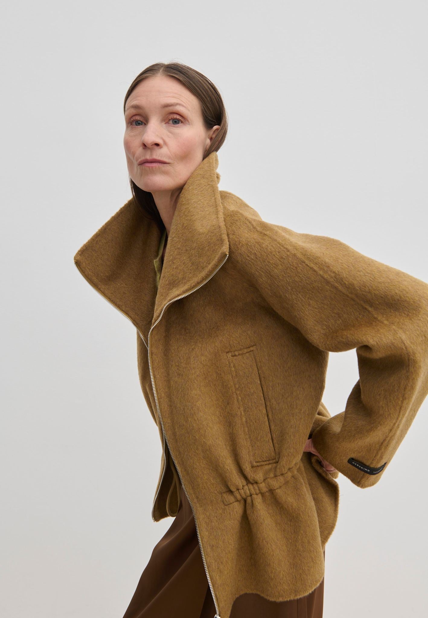 A woman wears a tan, funnel neck Oscar Wool Jacket by Herskind ApS with a zip front and a cinched waist.