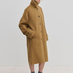 A person wearing a camel-colored Jacob Wool Coat by Herskind ApS, featuring a relaxed-fit, high collar, button-down front, and below-knee length.