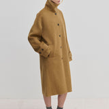 A person wearing a camel-colored Jacob Wool Coat by Herskind ApS, featuring a relaxed-fit, high collar, button-down front, and below-knee length.