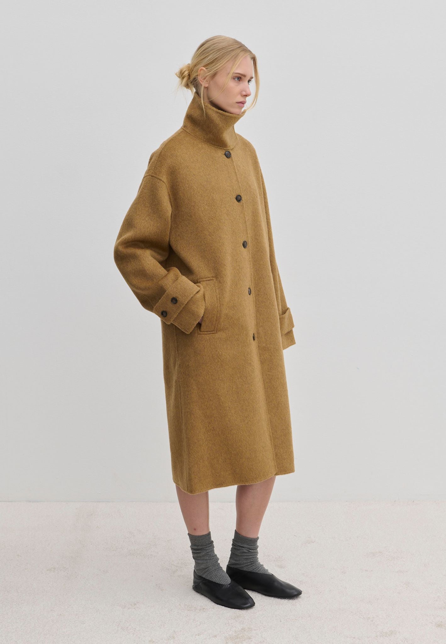 A person wearing a camel-colored Jacob Wool Coat by Herskind ApS, featuring a relaxed-fit, high collar, button-down front, and below-knee length.