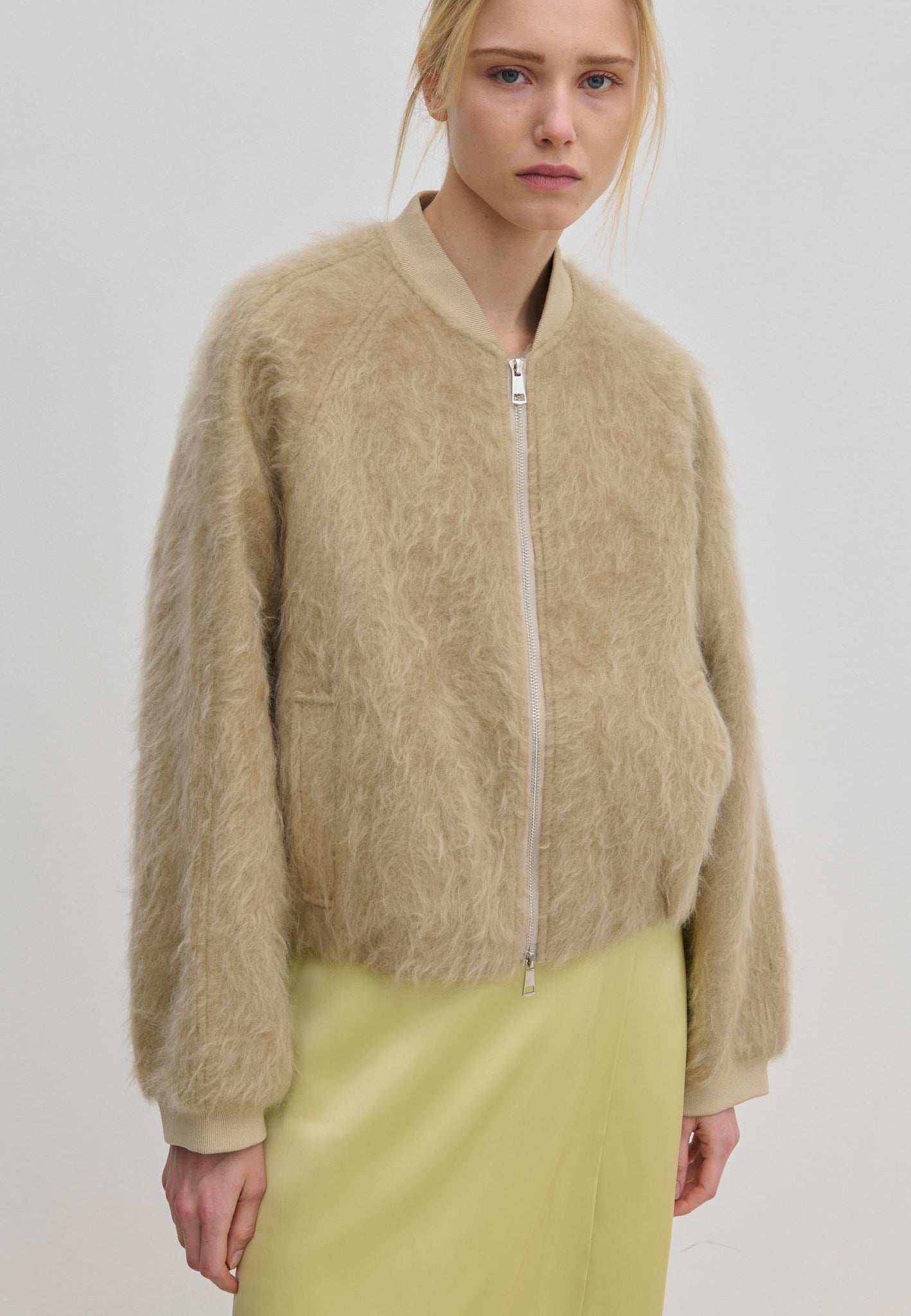 A relaxed fit, beige fuzzy Jimi Bomber Jacket by Herskind ApS with a front zipper, worn over a pale yellow skirt.