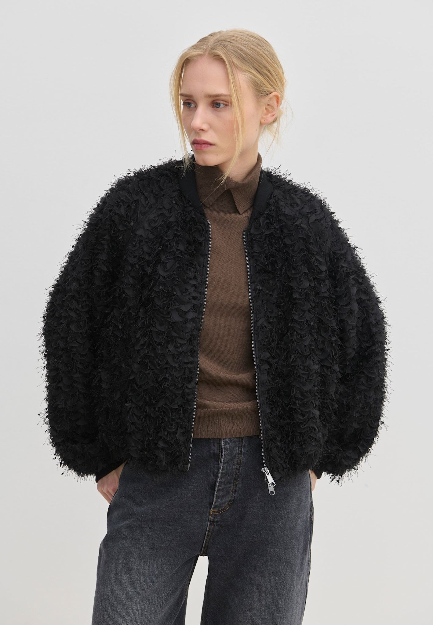The Jimi Bomber Jacket by Herskind ApS, a black bomber jacket with balloon sleeves and a textured, fringed surface, worn over a brown collared shirt and dark grey jeans.