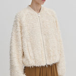 A person wears a cream-colored, relaxed-fit Jimi Bomber Jacket by Herskind ApS, featuring a textured, fringed finish and balloon sleeves.