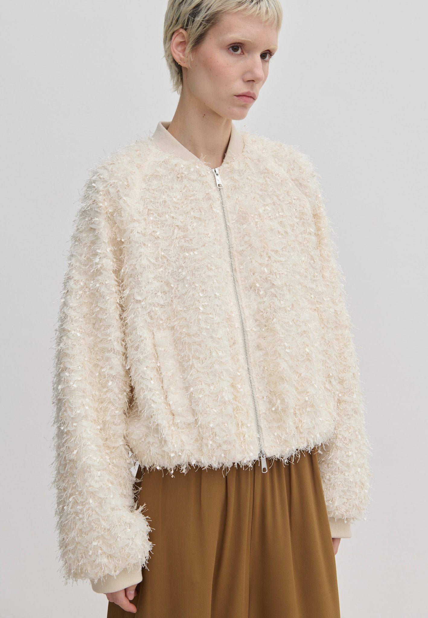 A person wears a cream-colored, relaxed-fit Jimi Bomber Jacket by Herskind ApS, featuring a textured, fringed finish and balloon sleeves.