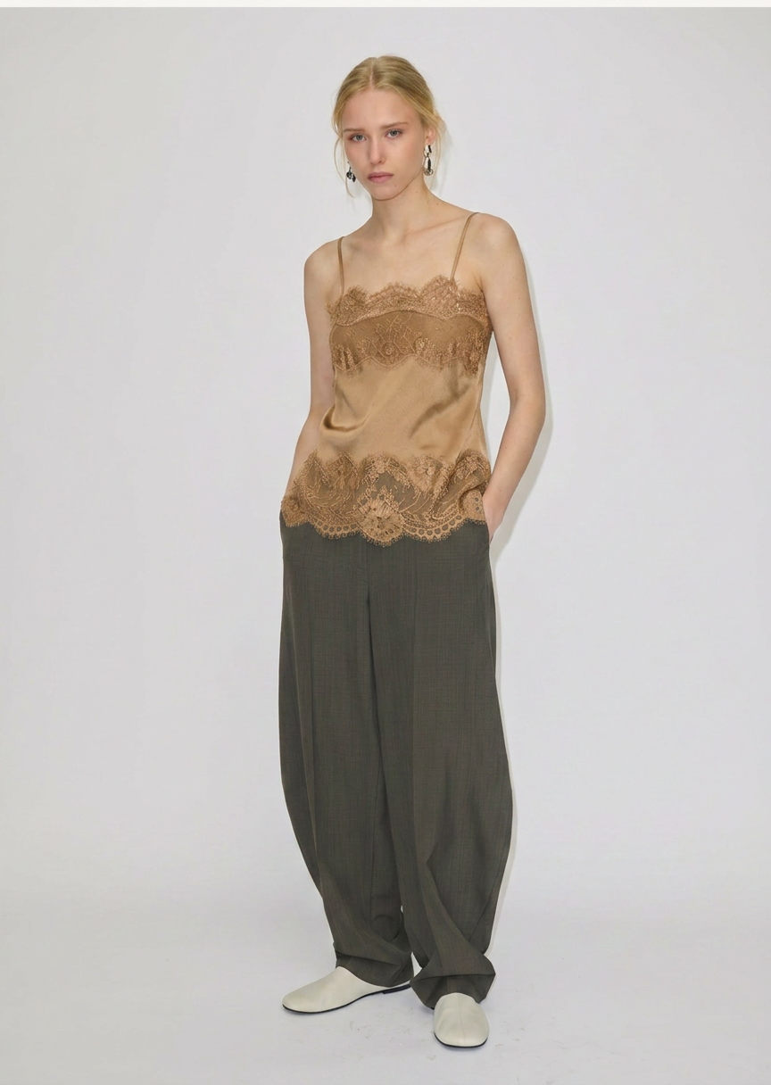 A model wears the tan Linnea Top with adjustable straps and lace trim at the neckline and hem, paired with loose-fitting olive trousers and white flat shoes.