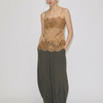 A model wears the tan Linnea Top with adjustable straps and lace trim at the neckline and hem, paired with loose-fitting olive trousers and white flat shoes.