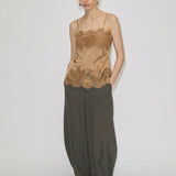 A model wears the tan Linnea Top with adjustable straps and lace trim at the neckline and hem, paired with loose-fitting olive trousers and white flat shoes.