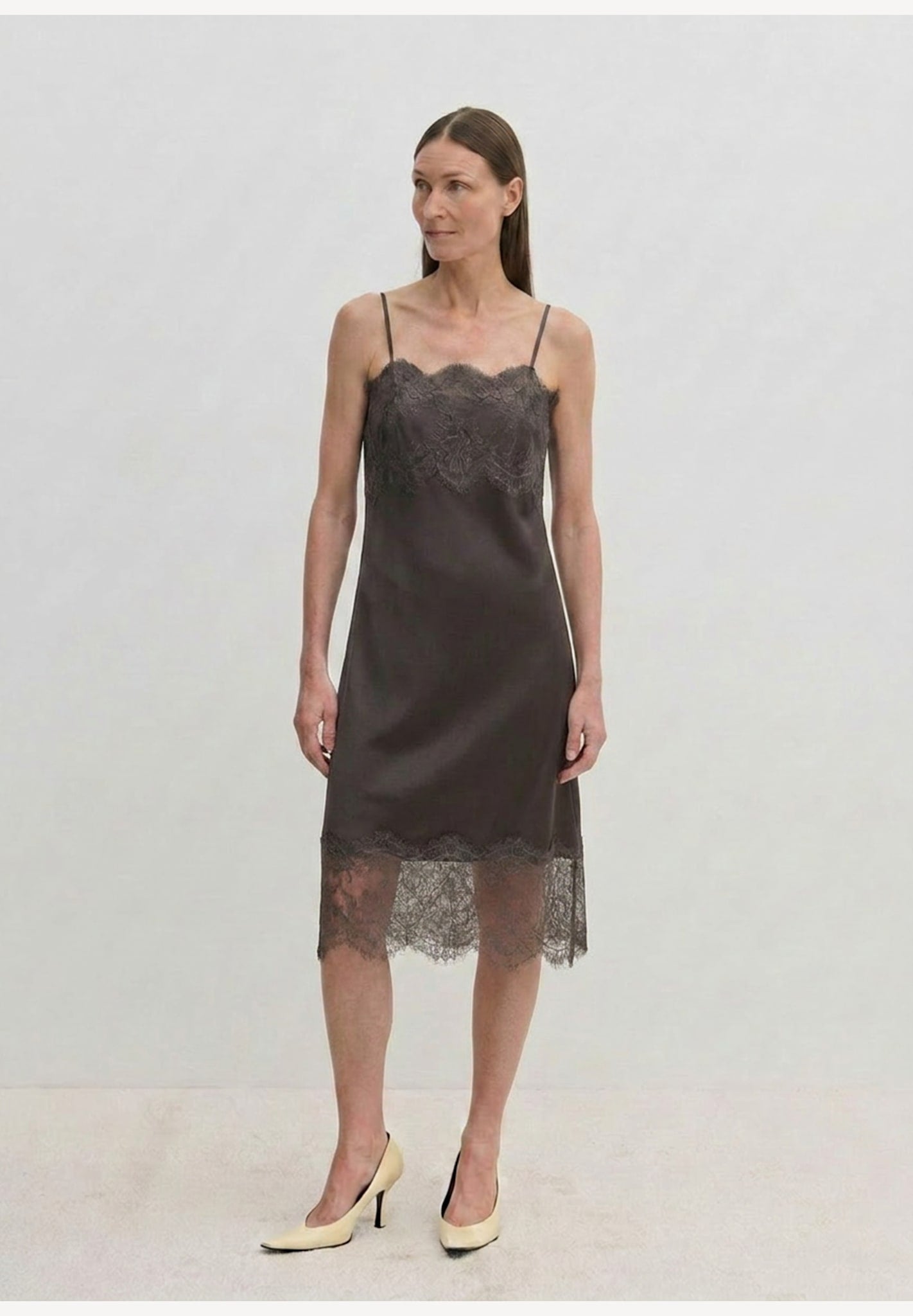 A person wears a dark midi-length Linus Dress by Herskind ApS with a lace-trimmed hem, paired with light-colored pointed-toe heels.