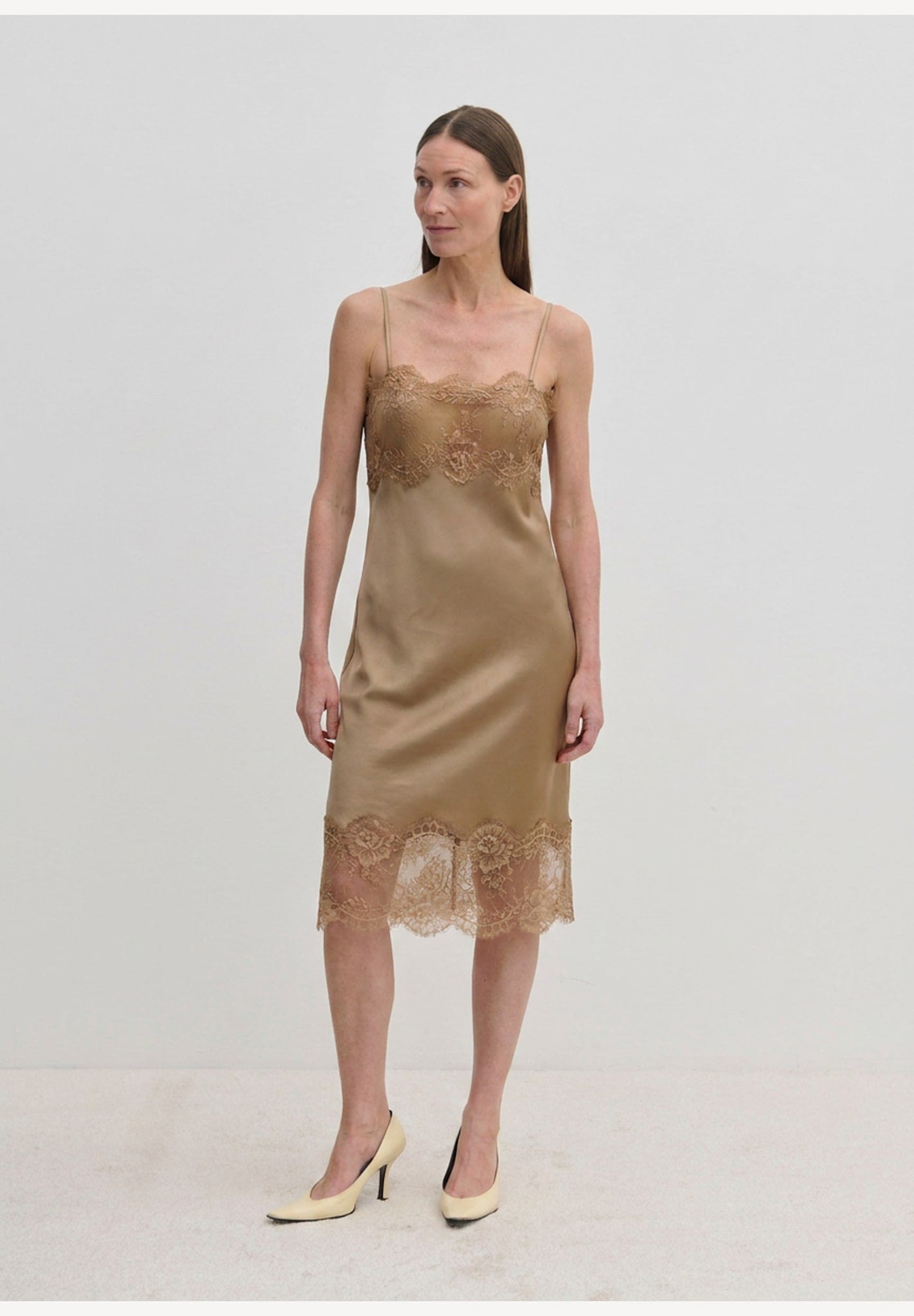 A tan midi-length Herskind ApS Linus Dress with lace-trimmed hem and neckline, worn with pointed-toe heels.