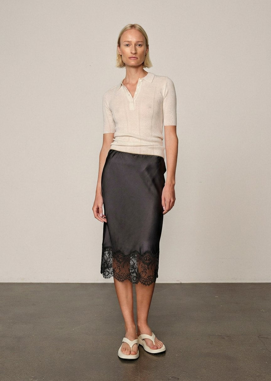 A woman wears a cream-colored polo shirt, a black satin Herbert Skirt midi slip skirt with a lace hem, and white thong sandals.