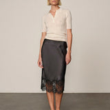 A woman wears a cream-colored polo shirt, a black satin Herbert Skirt midi slip skirt with a lace hem, and white thong sandals.