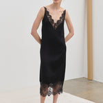 A model wearing the black Florence Dress by Herskind ApS, a midi slip dress featuring a V-neckline and hem trimmed in black lace.