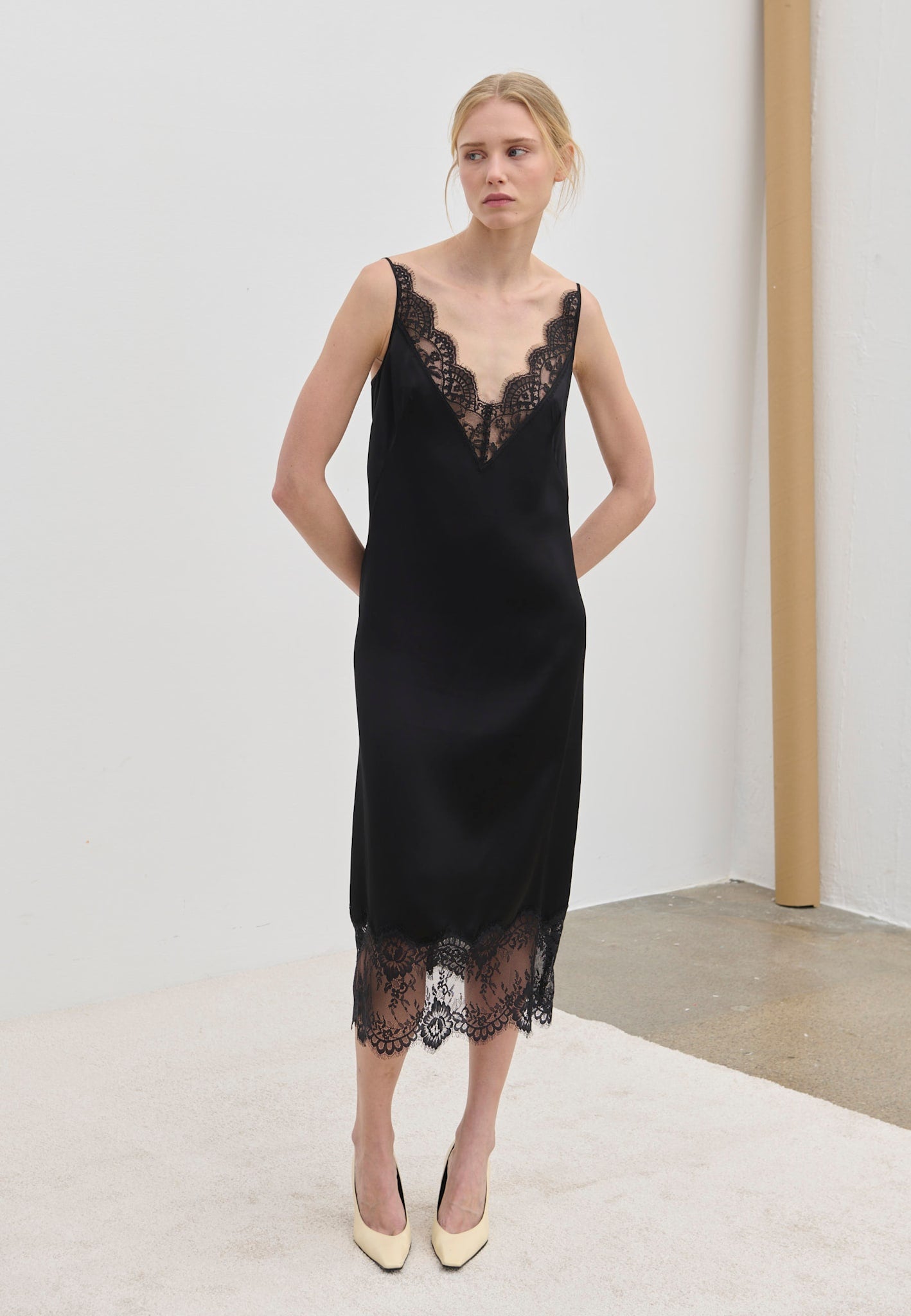 A model wearing the black Florence Dress by Herskind ApS, a midi slip dress featuring a V-neckline and hem trimmed in black lace.