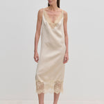 A woman wears a champagne-colored Florence Dress by Herskind ApS, a sleeveless midi slip dress with lace trim at the neckline and hem, paired with beige pointed-toe heels.