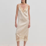 A woman wears a champagne-colored Florence Dress by Herskind ApS, a sleeveless midi slip dress with lace trim at the neckline and hem, paired with beige pointed-toe heels.