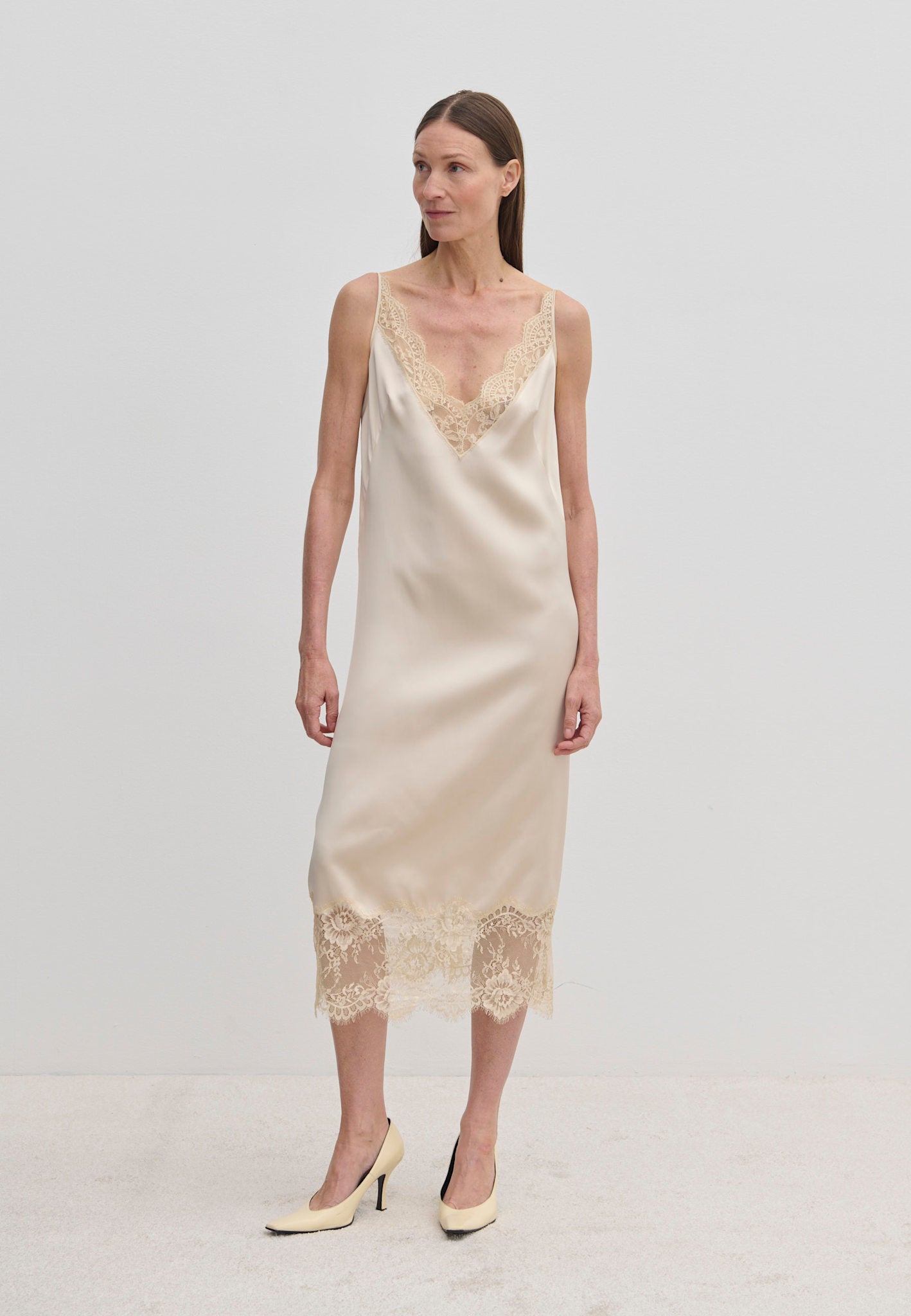 A woman wears a champagne-colored Florence Dress by Herskind ApS, a sleeveless midi slip dress with lace trim at the neckline and hem, paired with beige pointed-toe heels.