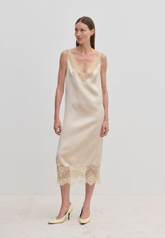 A woman wears a champagne-colored Florence Dress by Herskind ApS, a sleeveless midi slip dress with lace trim at the neckline and hem, paired with beige pointed-toe heels.