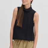 A woman wears a black, sleeveless Herskind ApS Loa Top with a draped funnel neckline and tan drawstring trousers.