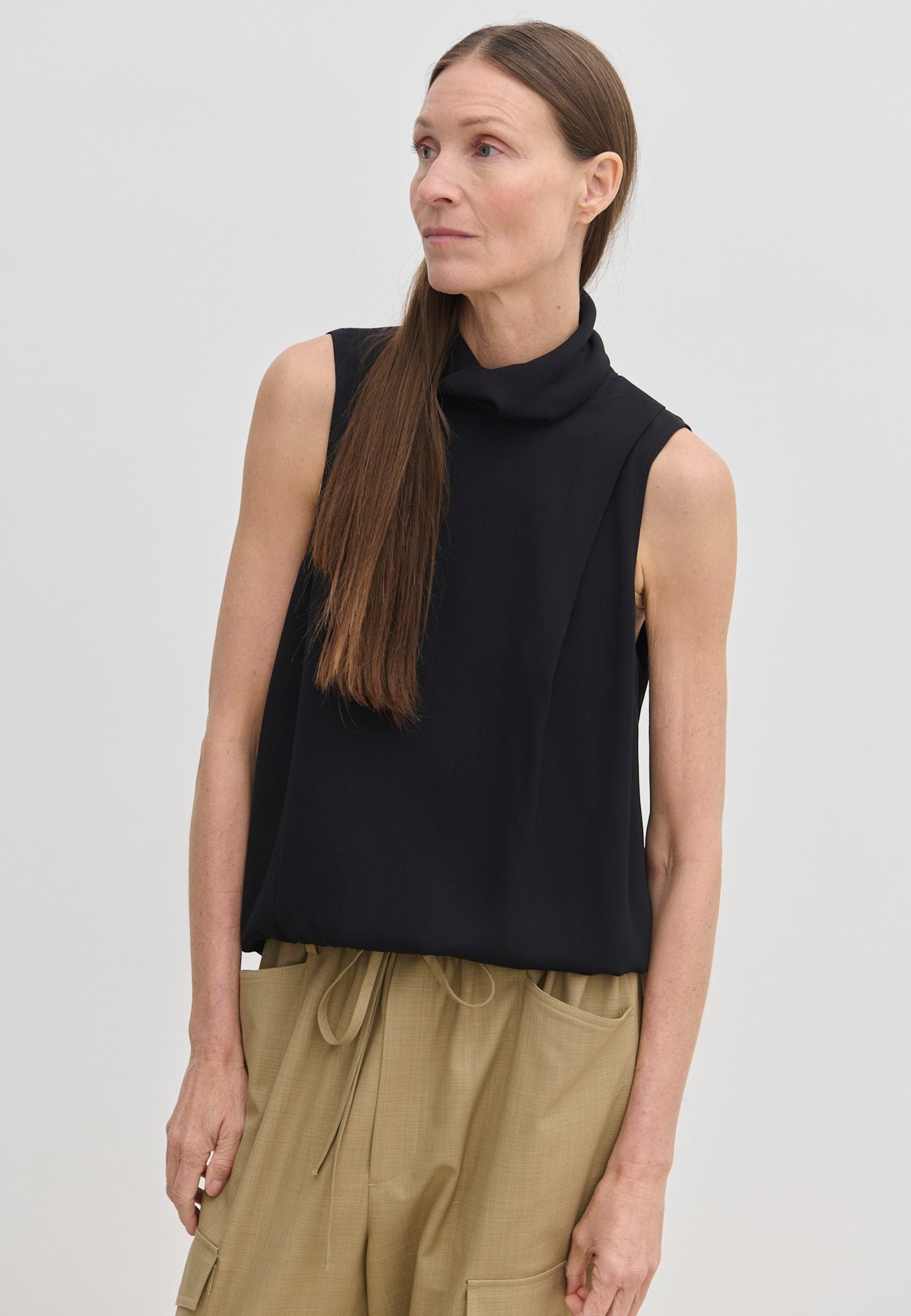 A woman wears a black, sleeveless Herskind ApS Loa Top with a draped funnel neckline and tan drawstring trousers.