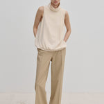 A model wearing a cream-colored Loa Top by Herskind ApS with a sleeveless, high funnel neck and a cinched hem, paired with tan trousers and cream-colored shoes.