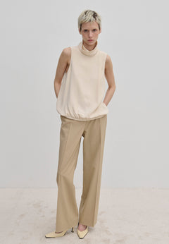 A model wearing a cream-colored Loa Top by Herskind ApS with a sleeveless, high funnel neck and a cinched hem, paired with tan trousers and cream-colored shoes.