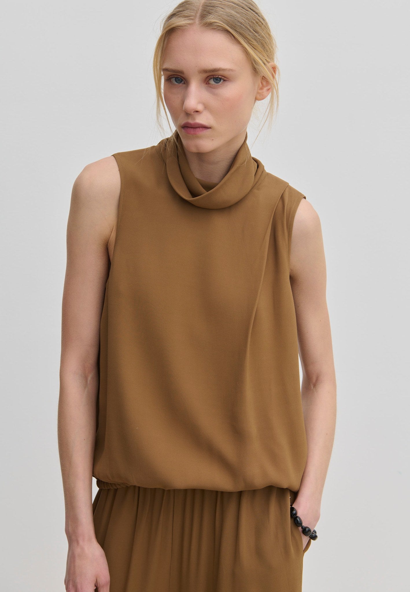 The Herskind ApS Loa Top is a sleeveless brown garment in a relaxed fit with a high, draped cowl neckline.