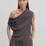 A person wearing a grey Scott Top by Herskind ApS featuring a one-shoulder, draped asymmetric neckline, paired with matching trousers.