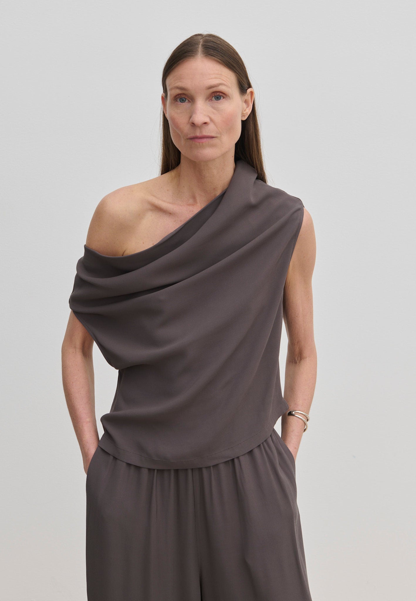 A person wearing a grey Scott Top by Herskind ApS featuring a one-shoulder, draped asymmetric neckline, paired with matching trousers.