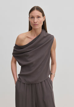 A person wearing a grey Scott Top by Herskind ApS featuring a one-shoulder, draped asymmetric neckline, paired with matching trousers.