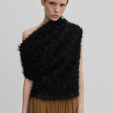 A model wears a black, textured, one-shoulder Scott Top by Herskind ApS, featuring a draped asymmetric neckline and a fringed appearance over a brown skirt.
