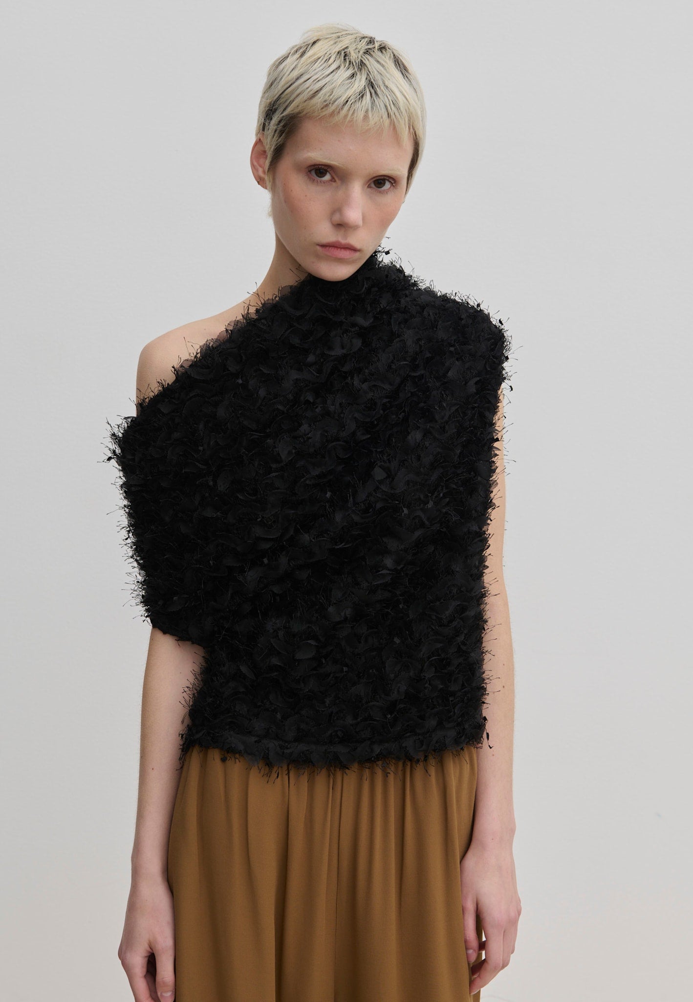 A model wears a black, textured, one-shoulder Scott Top by Herskind ApS, featuring a draped asymmetric neckline and a fringed appearance over a brown skirt.