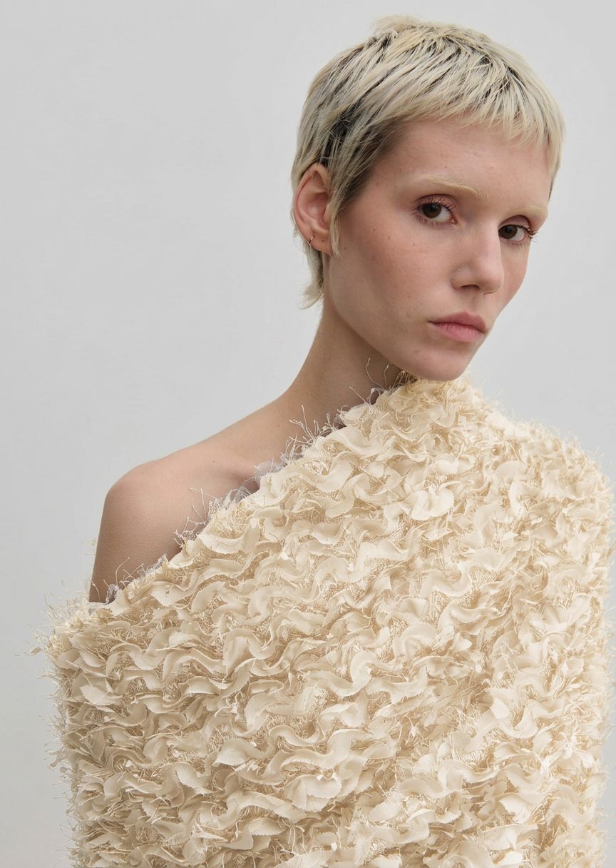 A person wearing the cream-colored Scott Top by Herskind ApS, featuring a relaxed-fit, off-the-shoulder design with layered, frayed ruffles and a draped asymmetric neckline.