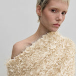 A person wearing the cream-colored Scott Top by Herskind ApS, featuring a relaxed-fit, off-the-shoulder design with layered, frayed ruffles and a draped asymmetric neckline.