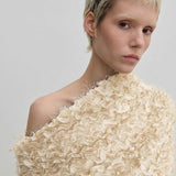 A person wearing the cream-colored Scott Top by Herskind ApS, featuring a relaxed-fit, off-the-shoulder design with layered, frayed ruffles and a draped asymmetric neckline.